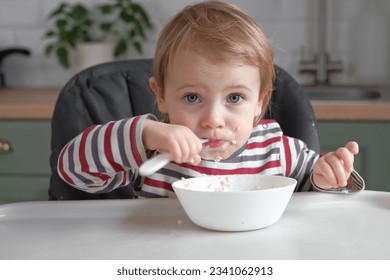 One year old hungry baby girl in striped casual clothes sits at white table in highchair and eats porridge herself with spoon. Blurred dining room background. Healthy eating for kids. Child nutrition - Powered by Shutterstock