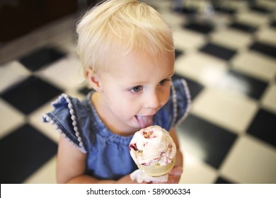 A One Year Old Little Girl Child Is Eating A Strawberry Vanilla Ice Cream Cone In An Old Fashioned Candy Shop With Black And White Checkered Floor Tiles.