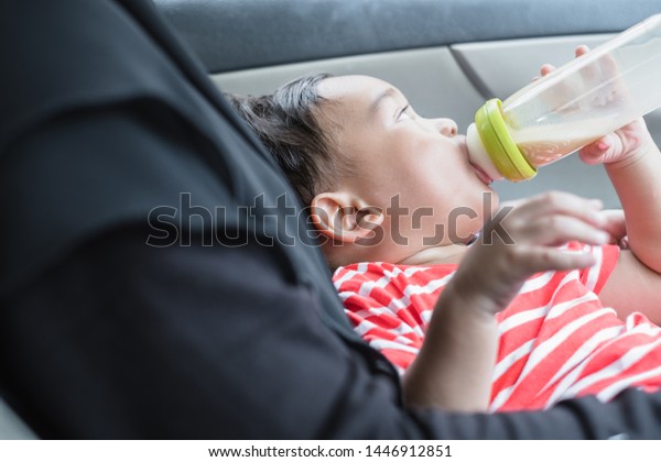 One year old Thai baby boy drink milk\
from a bottle in a car with mother\
hugging.