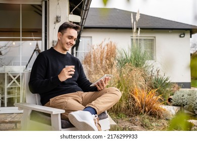 One young adult caucasian man sitting on the chair outdoor in front of his house in day while holding a cup of coffee and looking at digital tablet - Powered by Shutterstock