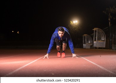 One Young Adult Man, Runner, Sprinter, Start Position, Running Track Red. Night Time Dark, Outdoors.