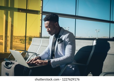 One Young And Attractive Black Businessman Is Seated In The Airport Terminal Working With A Laptop, And Smiling. He Is Carrying A Suitcase And Wearing Formal Clothes But Not A Suit.