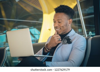 One Young And Attractive Black Businessman Is Seated In The Airport Terminal Working With A Laptop, And Smiling. He Is Carrying A Suitcase And Wearing Formal Clothes But Not A Suit.