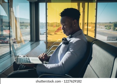 One Young And Attractive Black Businessman Is Seated In The Airport Terminal Working With A Laptop, And Smiling. He Is Carrying A Suitcase And Wearing Formal Clothes But Not A Suit.