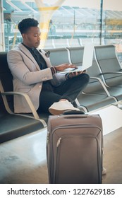 One Young And Attractive Black Businessman Is Seated In The Airport Terminal Working With A Laptop, And Smiling. He Is Carrying A Suitcase And Wearing Formal Clothes But Not A Suit.