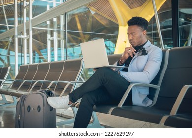 One Young And Attractive Black Businessman Is Seated In The Airport Terminal Working With A Laptop, And Smiling. He Is Carrying A Suitcase And Wearing Formal Clothes But Not A Suit.