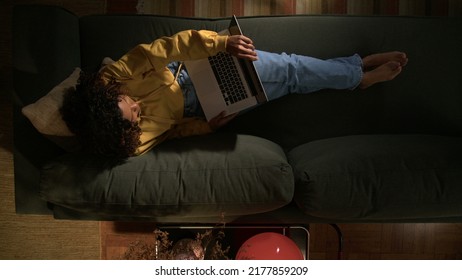 One Young Black Girl Lying Down On Couch Opening Laptop. Top View Angle Of Person Using Computer At Night