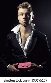 One Young Handsome Sensual Unshaven Bearded Man Looking At Camera With Popped Unbutton Collar White Shirt Black Jacket Keeping Pink Presentation Box With Bow In Studio On Black Background, Vertical