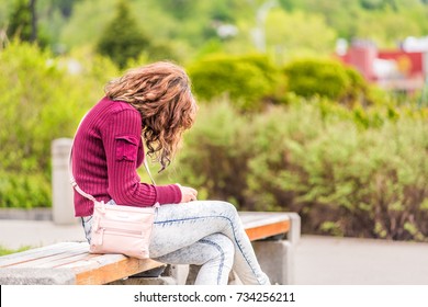 One young happy woman sitting on bench on sidewalk in green downtown city park in Saguenay, Canada, Quebec during summer looking at phone or reading book