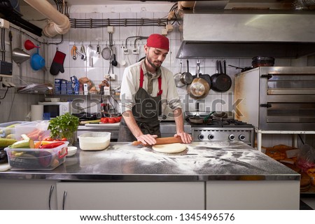 One young man, 20-29 years old, preparing pizza dough in commerical kitchen, a table indoors.