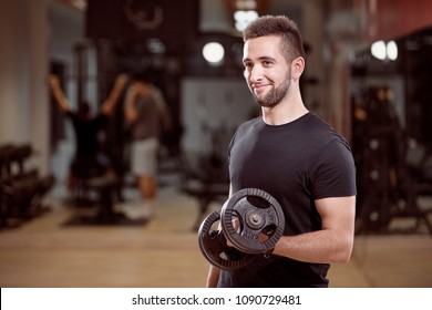 One Young Man, Ordinary Average Looking, One Arm Dumbell Exercise, Smirking, In Gym. Unrecognizable Group Of People Behind.