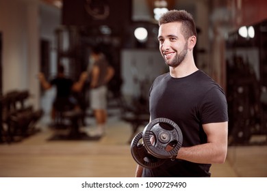 One Young Man, Ordinary Average Looking, One Arm Dumbell Exercise, Smiling, In Gym. Unrecognizable Group Of People Behind.