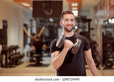 One Young Man, Ordinary Average Looking, One Arm Dumbell Exercise, Smirking, In Gym. Unrecognizable Group Of People Behind.