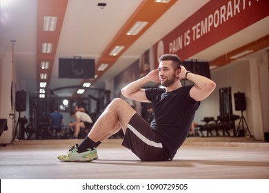 One Young Man, Ordinary Average Man, Exercise Abs, Sideways, Sitting On Floor, In Gym. Unrecognizable People Behind (out Of Focus).