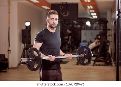 One Young Man Posing, Ordinary Average Looking, Holding Barbell With Weights, Exercise In Gym. Unrecognizable Person Behind (out Of Focus).