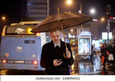 one young man, standing outdoors on a street using his phone, and holding an umbrella. Waiting at a city bus station.