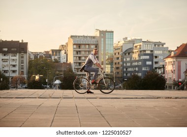 One Young Smiling Man, 20-29 Years Old,  Wearing Hipster Suit, Smart Casual, Sitting On Old City Bike. City Buildings Panorama Behind In Background. Full Lenght Shot.