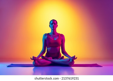 One young sportive woman sitting in lotus yoga pose on mat and doing meditation session isolated over gradient pink and yellow background in neon. Concept of youth, sport, health, fitness, wellness - Powered by Shutterstock