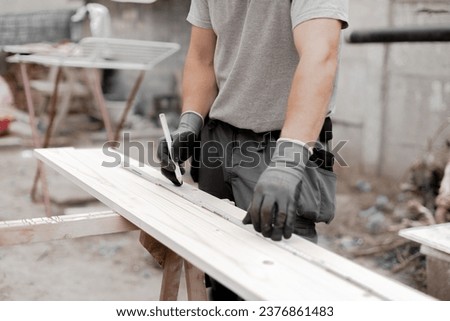 One young unrecognizable Caucasian man in gray gloves holds a construction measuring tape and draws on a board with a pencil, standing in the backyard of a house on a summer day, side view close-up.