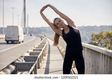 One Young Woman, 20-29 Years Old, Stretching On Bridge, Van In Background. Black Sport Clothes, Summer Day.