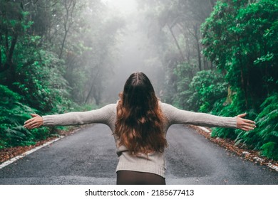 One young woman enjoying and discovering alone forest walking in the middle of the road with green trees around her. Girl opening arms and looking up feeling free. Freedom concept.