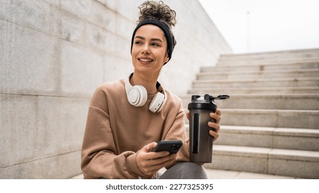 One young woman with headphones hold supplement shaker bottle and smartphone mobile phone sporty caucasian female sit take a brake for training generation z healthy lifestyle concept copy space - Powered by Shutterstock