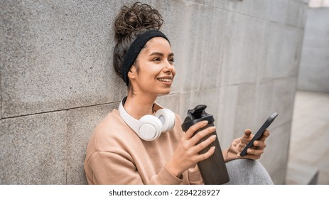 One young woman with headphones hold supplement shaker bottle and smartphone mobile phone sporty caucasian female sit take a brake for training generation z healthy lifestyle concept copy space - Shutterstock ID 2284228927