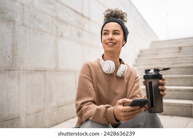 One young woman with headphones hold supplement shaker bottle and smartphone mobile phone sporty caucasian female sit take a brake for training generation z healthy lifestyle concept copy space - Powered by Shutterstock