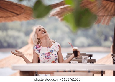 One Young Woman Laughing, 25 Years Old, Looking Up Above, Sitting In Beach Cafe, Drinking Coffee From Cup. Tables Leaves Sunshades.