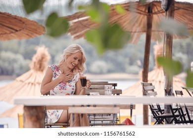 One Young Woman Laughing, 25 Years Old, Sitting In Beach Cafe, Drinking Coffee From Cup. Tables Leaves Sunshades.