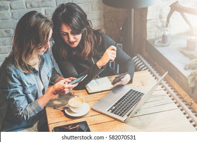 One-on-one meeting. Two young business women sitting at table in cafe. Girl shows her friend image on screen of smartphone. Second girl holding smartphone. On table is laptop, notebook and cup of coffee.