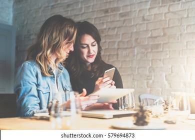 One-on-one meeting.Two young business women sitting at table in cafe.Girl shows her friend image on screen of smartphone.Second girl holding digital tablet.On table is closed notebook.Meeting friends.