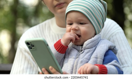 A One-year-old Boy Sits In His Mother's Arms In The Park And Watches A Cartoon On The Phone In The Park Close-up. Happy Child Waving Hands While Watching Cartoons On Smartphone.