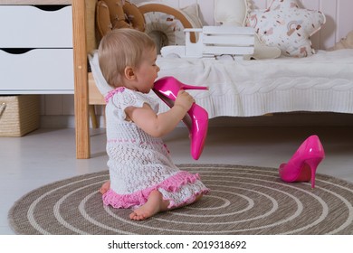 A One-year-old Girl In A White And Pink Openwork Dress Licks Her Mother's Women's Hot Pink High-heeled Pumps While Sitting On The Floor In The Children's Room