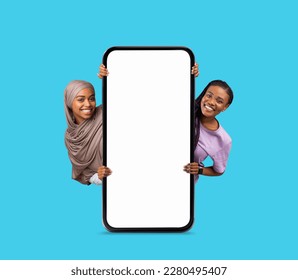 Online Advertisement. Two Happy Females Holding Big Blank Smartphone With White Screen, Smiling African American Ladies Demonstrating Copy Space For Mobile Offer, Standing On Blue Background, Mockup - Powered by Shutterstock