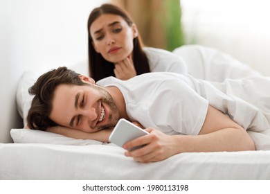 Online Affair. Wife Spying While Cheating Husband Texting On Cellphone Having Flirty Chat Conversation With Lover Lying In Bed At Home. Jealous Woman Checking Her Boyfriend's Phone. Selective Focus