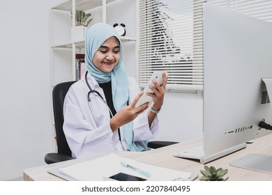 Online Appointment With Doctor Concept. Muslim Woman Doctor Working In Clinic, Sitting At Workdesk And Using Digital Tablet, Making Online Appointment With Patient Or Training