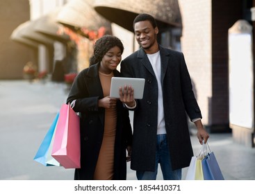 Online banking application, ad of sales and promotions in stores. Happy african american young couple with many multicolored shopping bags looking at tablet, walking in city near city mall, free space