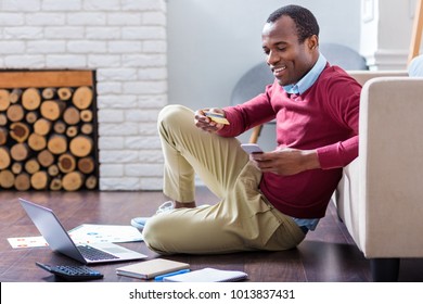 Online Banking. Happy Smart Nice Man Holding His Smartphone And Looking At The Credit Card While Making An Online Payment