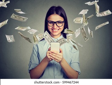 Online banking money transfer, e-commerce concept. Happy young woman in glasses using smartphone with dollar bills flying away from screen isolated on gray wall background.
