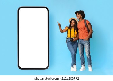 Online booking concept. Happy young black couple with camera and backpack tourists hugging and pointing at big smartphone with white empty screen, mockup, blue studio background, full length