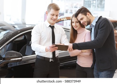 Online Booklet. Handsome Young Man Embracing His Beautiful Girlfriend Looking At The Digital Tablet Car Salesman Is Showing Him At The Car Dealership Couple Buying A Car Service Professionalism Choice