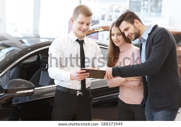 Online booklet. Handsome young man embracing his\
beautiful girlfriend looking at the digital tablet car salesman is\
showing him at the car dealership couple buying a car service\
professionalism choice