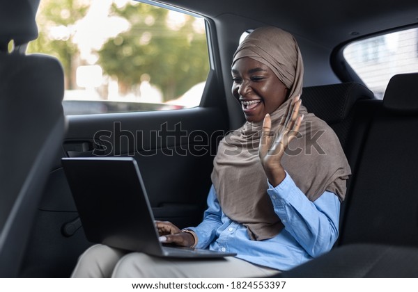 Online\
Business Meeting. Smiling african muslim businesswoman making video\
call with laptop in car, waving hand at camera, greeting partners,\
going to office, sitting on backseat with\
computer