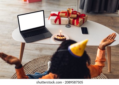 Online Celebration. Rear-View Of Black Female At Laptop With Blank Screen Celebrating Her Birthday Or New Year Wearing Festive Hat, Having B-Day Cake And Wrapped Gifts On Table At Home
