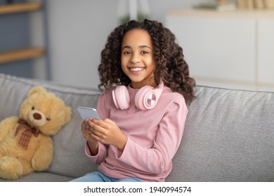 Online Chat. Cheerful Black Girl Using Celphone With Headphones On Neck