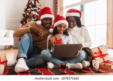 Online Christmas Party. Happy Black Family In Red Santa Claus Hat Using Laptop Pc Making Video Call Celebrating Xmas Distantly Sitting On Floor At Home, Holding Showing Gifts. New Year Holiday Concept - Powered by Shutterstock