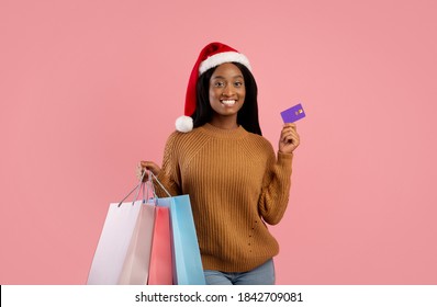 Online Christmas sale concept. Happy black woman in Santa hat holding shopping bags and credit card on pink studio background. Positive African American lady with packed Xmas purchases and bank card - Powered by Shutterstock