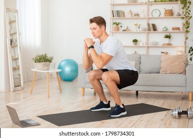 Online Coach For Home Workout. Handsome Young Man In Sportswear With Fitness Tracker, Squats And Looks At Laptop In Living Room Interior, Free Space