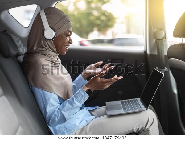 Online Communication. Black Muslim Woman Having\
Video Conference Via Laptop In Car, Sitting On Backseazt Of\
Automobile, Making Video Call With Computer And Headset, Talking At\
Device Camera, Side View